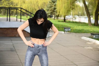 Bahar havasında beyaz tenli esmer bir kızın ince bir figürle poz verdiği portresi. Model, güzel havada bir şehir parkının arka planına karşı duruyor..