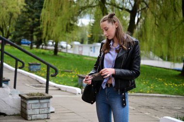 Şehrin dışındaki bahar parkında poz veren bir model. Siyah ceketli ve kot pantolonlu, koruyucu maskeli genç bir kızın fotoğrafı..