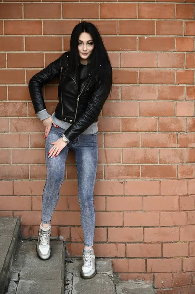 Full-length portrait in spring weather outdoors, caucasian brunette girl in jeans and a black jacket with a great figure. Model posing against the backdrop of a building wall.