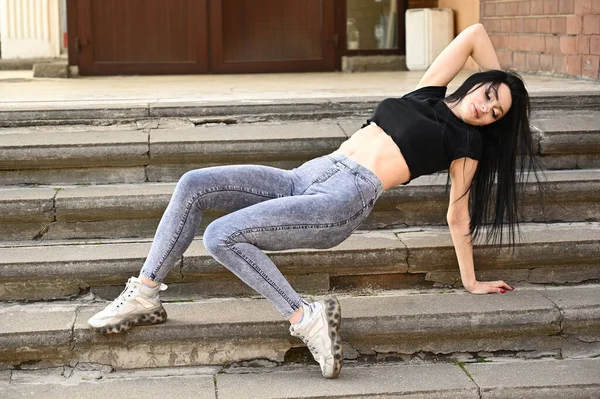 Bahar havasında açık havada tam boy portre, kot pantolon ve siyah tişörtlü beyaz tenli kız. Binanın merdivenlerinde bir model oturuyor..