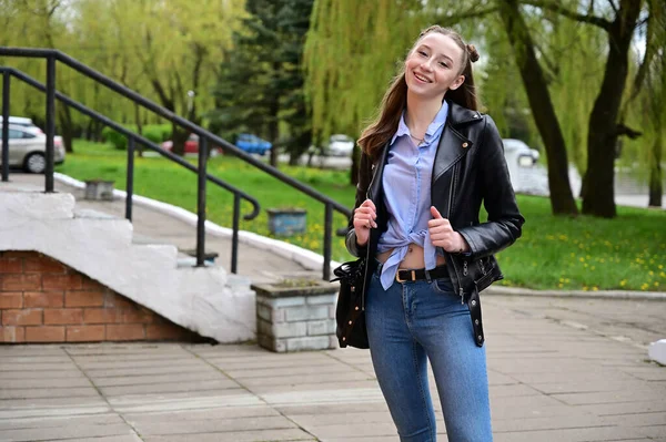一个穿着黑色夹克和牛仔裤笑着的年轻漂亮姑娘的画像 模特儿站在城外的春天公园里 — 图库照片