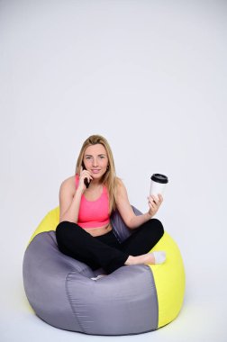 İnce, beyaz bir model gülümseyerek poz veriyor. Beyaz arka planda spor üniformalı güzel bir kızın portresi elinde bir fincan kahveyle yumuşak bir sandalyede oturuyor..