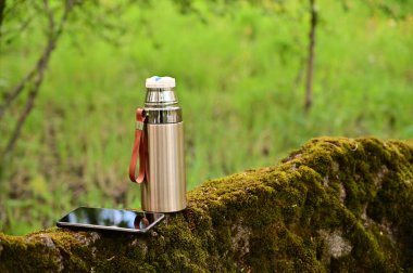 Yazın açık havada ormanda bir termos ve akıllı telefonun fotoğrafı. Turist tatili kavramı.
