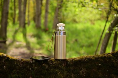 Yazın açık havada ormanda bir termos ve akıllı telefonun fotoğrafı. Turist tatili kavramı.