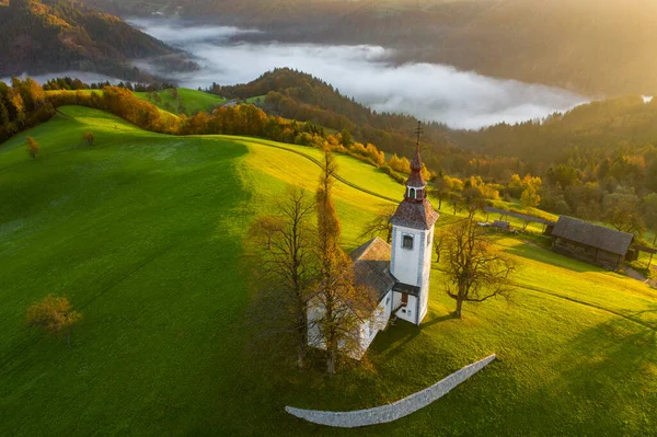 Vista aérea das colinas, floresta colorida no nevoeiro e da igreja de Sv Tomaz. Nascer do sol na Eslovénia no Outono — Fotografia de Stock