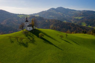 Tepelerin havadan görünüşü, renkli ormanlar ve Sv Tomaz kilisesi. Slovenya 'da sonbaharda gün batımı