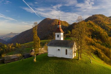 Tepelerin havadan görünüşü, renkli ormanlar ve Sv Tomaz kilisesi. Slovenya 'da sonbaharda gün batımı