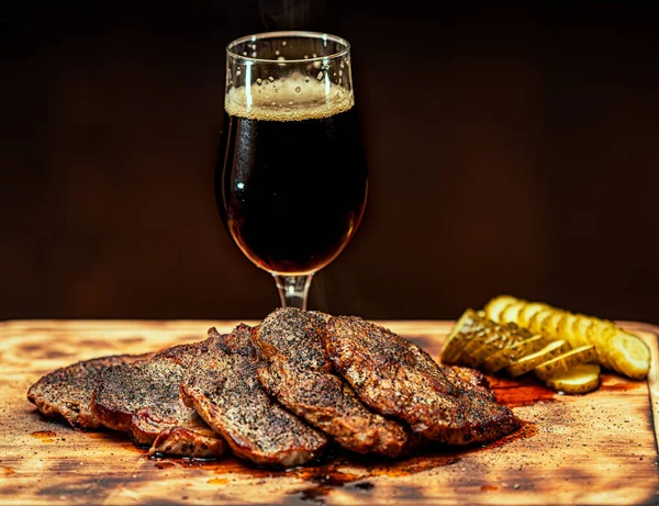 黒を基調とした木のテーブルの上に肉前菜と軽い泡ビールのガラス — ストック写真