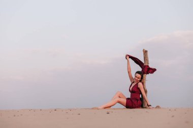 Çölde, rüzgarda, yaşlı bir ağacın yanında kırmızı bir elbiseyle oturan kız.