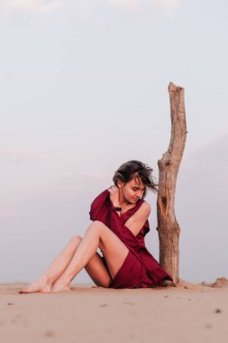 Çölde, rüzgarda, yaşlı bir ağacın yanında kırmızı bir elbiseyle oturan kız.