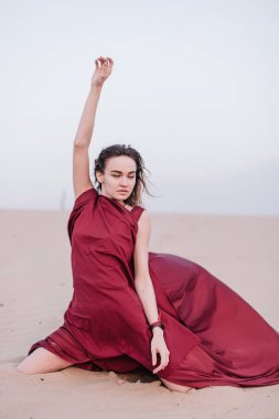 Kırmızı elbiseli kız kumda oturuyor.