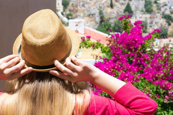 若いですブロンドの女の子とともに波状の髪でわら帽子は開花オレンジ色の花やギリシャ島の小さなカラフルな家で見ていますシミ島のDdekanisos諸島 — ストック写真