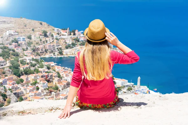 Hasır şapkalı dalgalı saçlı genç sarışın kız dağın tepesinde oturuyor ve Dodekanisos takımadalarında Yunan adası Symi'de küçük renkli evleri ve Akdeniz'i izliyor. 