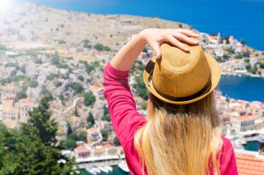 Güneş gözlüklü dalgalı saçlı genç kız dağın tepesinde oturuyor ve Dodekanisos takımadalarındaki Yunan adası Symi 'deki küçük renkli evleri ve Akdeniz' i izliyor.