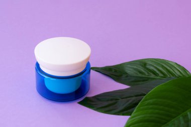 Mavi kavanozda yumuşak beyaz krema ve mor arka planda parlak yeşil yapraklar. Evde spa tedavisi ve prosedür.