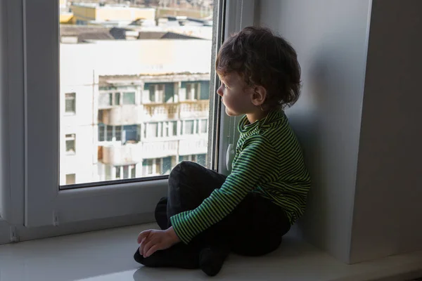 quarantine during a coronavirus pandemic. . The child stands on the windowsill and looks at the empty city through the window. Dreams to go out and breathe fresh air