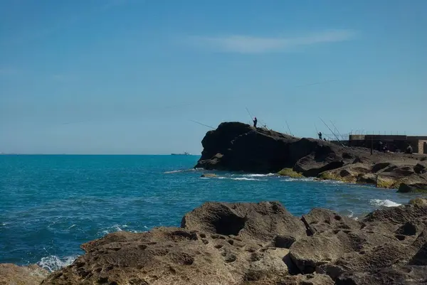 Yüksek bir kayanın üzerinde elinde oltayla bir balıkçı. Deniz kıyısında..