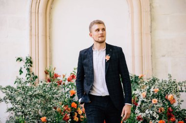 Takım elbiseli damat, açık hava düğün töreni için mercan renginde taze çiçeklerin alışılmadık bir kompozisyonu karşısında poz veriyor.