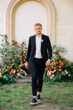 Takım elbiseli damat, açık hava düğün töreni için mercan renginde taze çiçeklerin alışılmadık bir kompozisyonu karşısında poz veriyor.