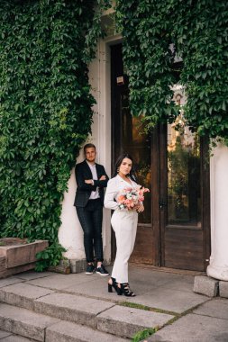 Gelin ve damat beyaz bir takım elbiseyle sarmaşıkla kaplanmış bir binanın arka planında poz veriyorlar.