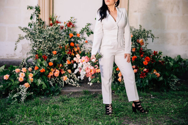original bride in a white trouser suit posing against the background of an unusual composition of fresh flowers in coral color for an outdoor wedding ceremony