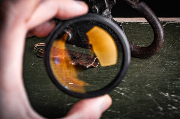 обрезанное изображение человеческой руки с лупой, ржавым замком и ключом на столе
