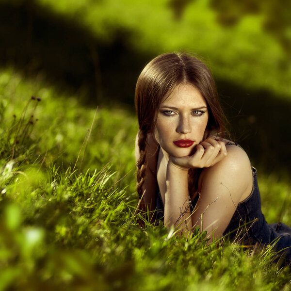 Front view toned portrait of a beautiful young woman with a thoughtful look and freckles, lying at green grass during a sunny spring day