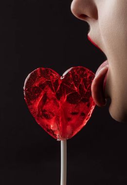 cropped image of woman licking red heart shaped lollipop isolated on black, valentines day concept clipart