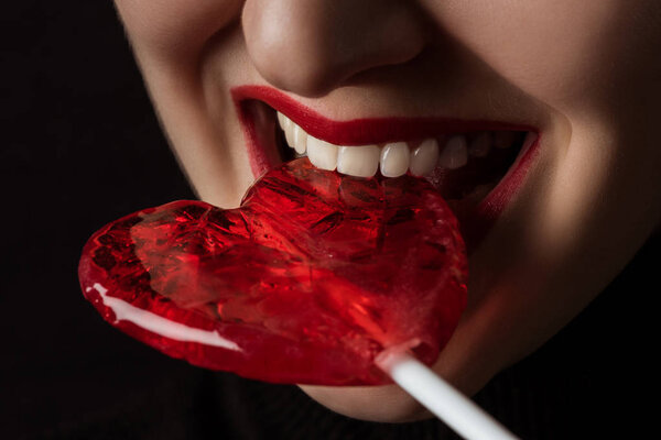 cropped image of woman biting heart shaped lollipop isolated on black, valentines day concept 