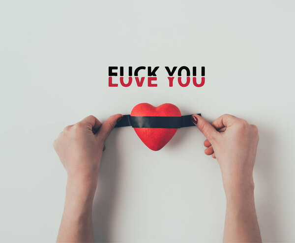 Cropped image of woman putting insulating tape on red heart with words love you fuck you on white