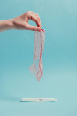cropped shot of woman holding ripped condom in hand and pregnancy test isolated on blue clipart