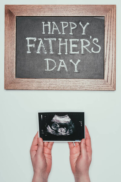 cropped shot of woman with ultrasound scan in hands and happy fathers day blackboard isolated on grey