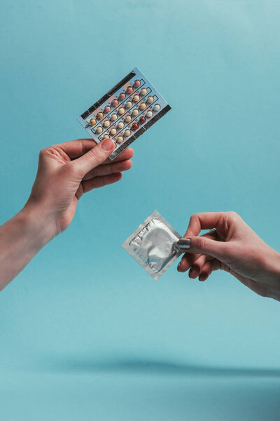 partial view of women holding condom and contraceptive pills in hands isolated on blue