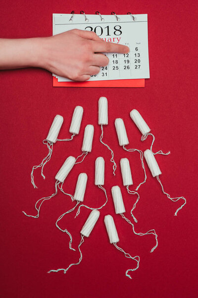 cropped shot of woman pointing at date in calendar with menstrual tampons around isolated on red