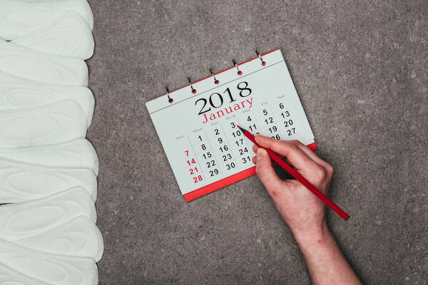 обрезанный снимок женщины, указывающей на дату в календаре с менструальными прокладками вокруг на серой поверхности
