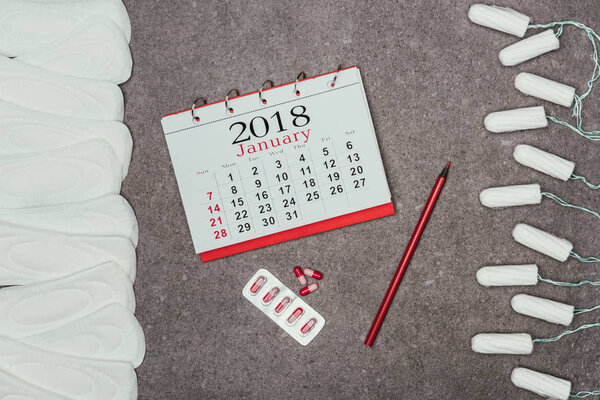 top view of arranged menstrual pads and tampons, calendar and pills on grey surface