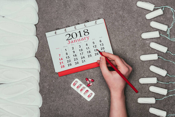 cropped shot of woman pointing at date in calendar with pills, menstrual pads and tampons around on grey surface