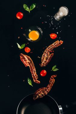 close up view of pieces of bacon, cherry tomatoes and raw egg yolk falling on frying pan isolated on black