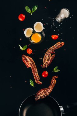 close up view of pieces of bacon, cherry tomatoes and raw egg yolk falling on frying pan isolated on black clipart