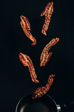 close up view of pieces of bacon falling on frying pan isolated on black