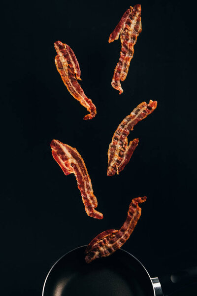 close up view of pieces of bacon falling on frying pan isolated on black