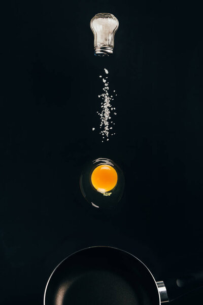 close up view of raw egg yolk and salt falling on frying pan isolated on black
