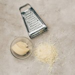 Grater and piece of cheese in bowl on light background