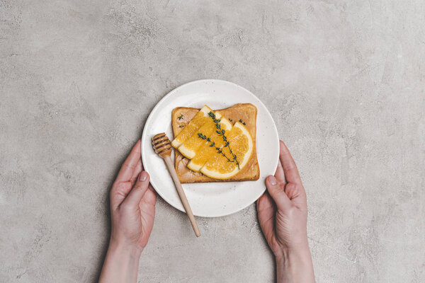 partial top view of person holding plate with fresh healthy sandwich with honey and orange slices on grey