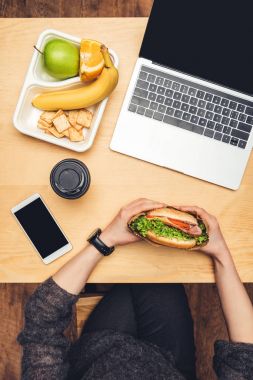 cropped image of woman eating burger at table with fruits and gadgets clipart