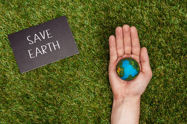 cropped image of man holding earth model in hand with sign save earth on green grass, earth day concept