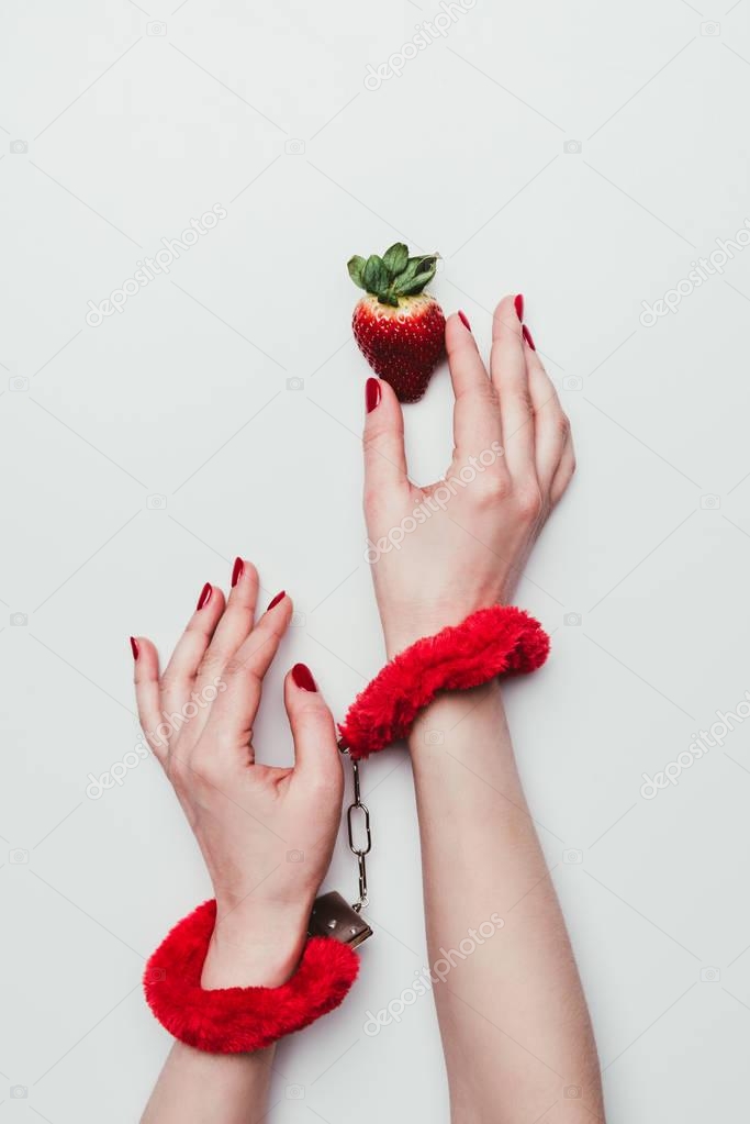 Female hands in red fluffy handcuffs reaching for strawberry isolated on white