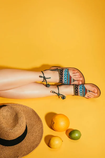 cropped shot of citrus fruits, woman feet in stylish sandals and straw hat on yellow background