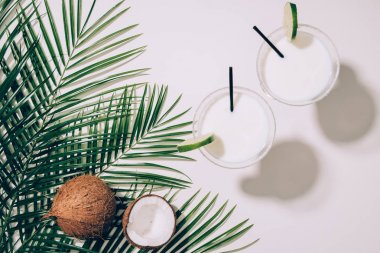 top view of organic coconuts, green palm leaves and coconut cocktails in glasses with drinking straws on white  clipart