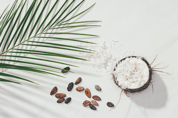 top view of half of coconut with shavings and cocoa beans with green palm leaves on white